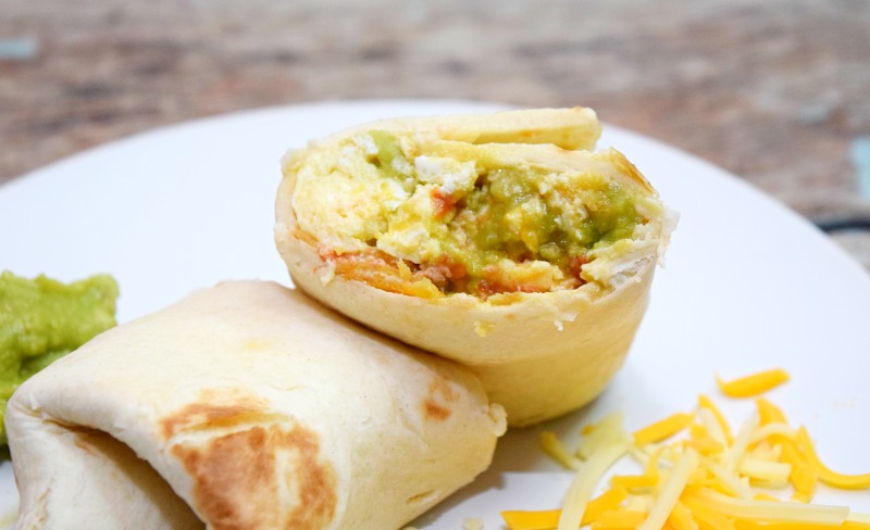 A close up shot of migas in a flour tortilla surrounded by guacamole and cheese. 