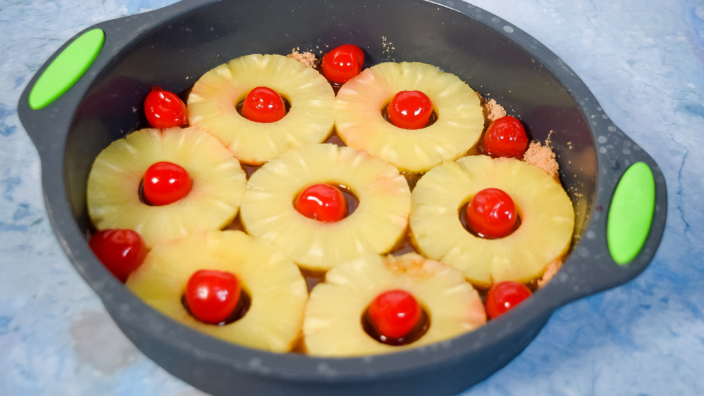 Pineapple rings and red cherries and brown sugar in a gray silicone cake pan. 