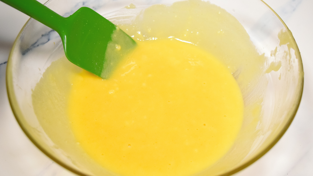 A glass bowl with melted butter mixed with yellow cake mix with a green spatula on a marble background. 