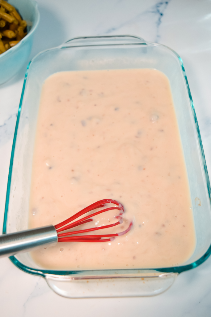 A clear casserole dish with a red whisk mixing up mushroom soup and milk.