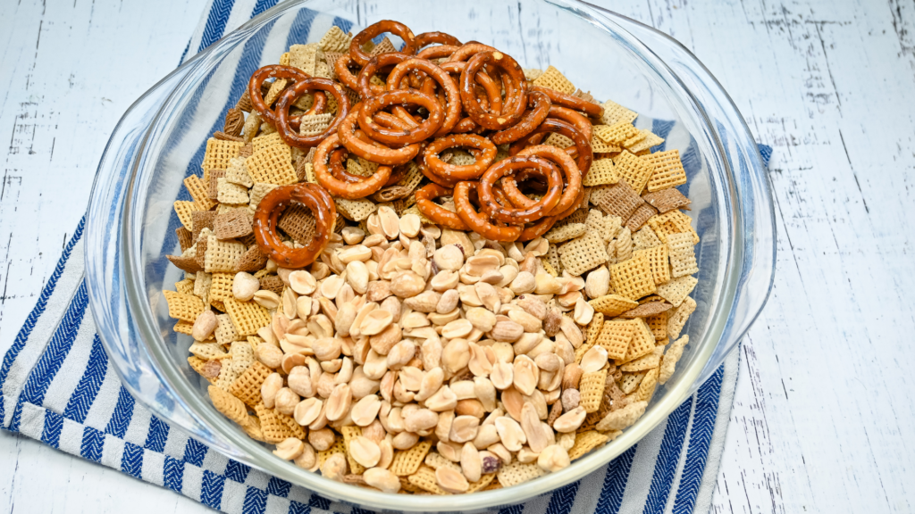 The cereal, nuts, and pretzels all in a clear glass bowl on a blue striped towel ready to make Microwave Chex Mix