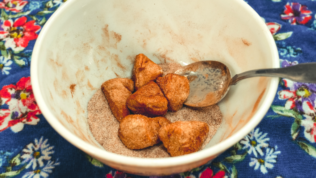 A bowl on a blue floral towel with cinnamon and sugar and cut pieces of biscuit to be coated. 