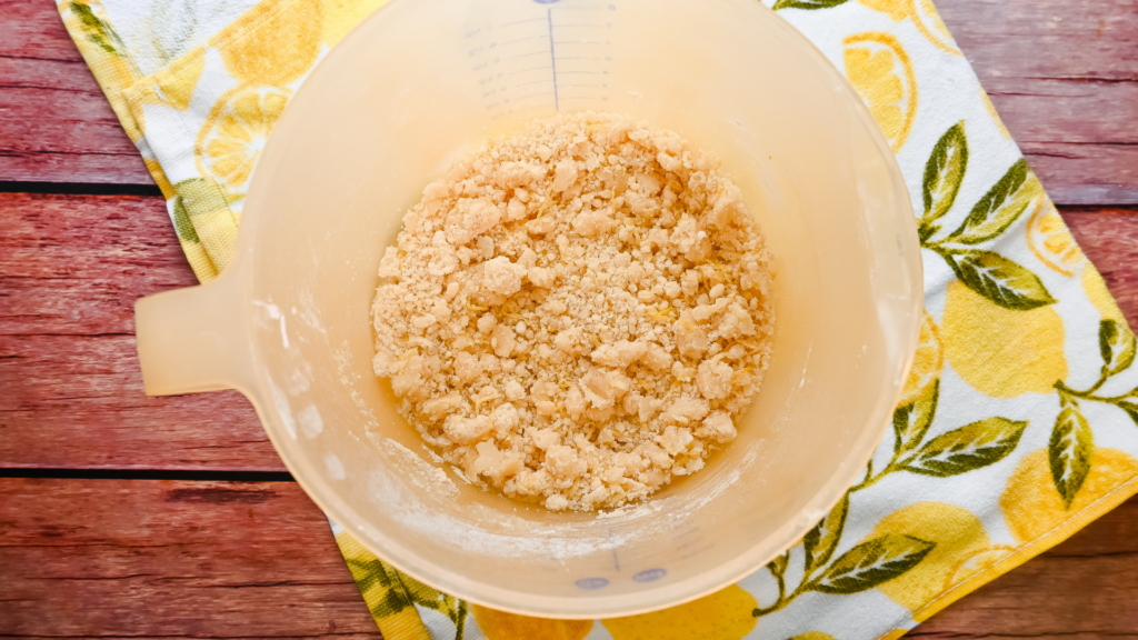 A platstic bowl full of a crumbly crust mixture on a wooden background with a lemon towel