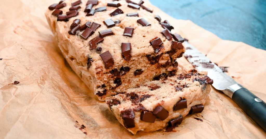 A finished loaf of Chocolate Chip Banana Bread in the Microwave on brown parchment paper with a knife with one slice cut. 