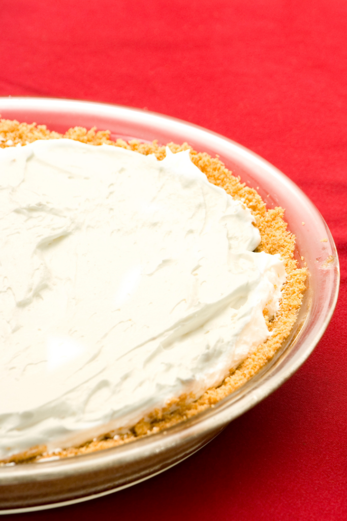 A close up of no bake cheesecake in a graham cracker crust in a glass pie plate on a red background. 
