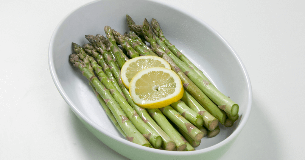 Adding asparagus to an white oval microwave-safe baking dish with two slices of lemon before cooking in the microwave