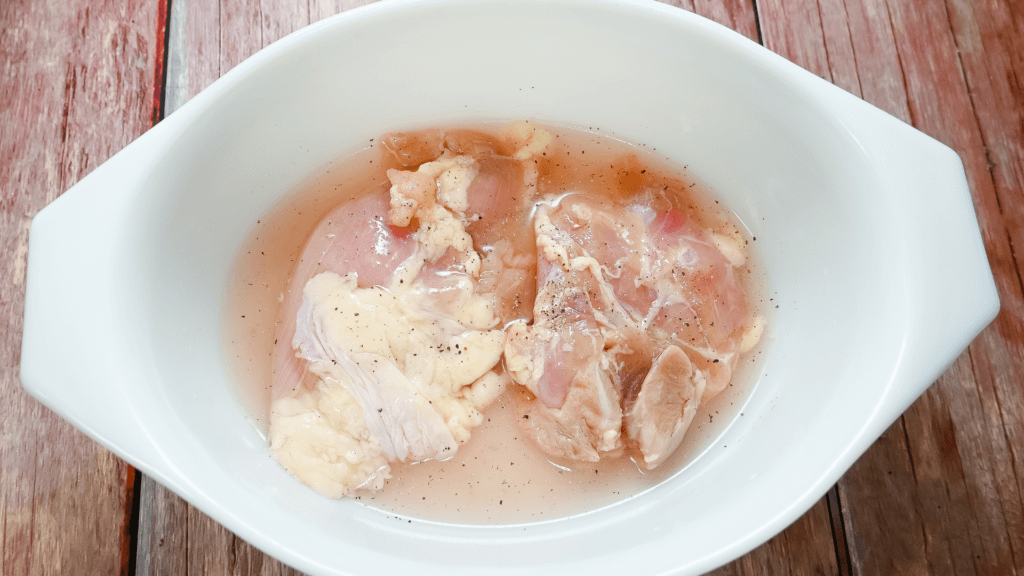 Raw chicken thighs seasoned and in water in a white bowl ready to be cooked