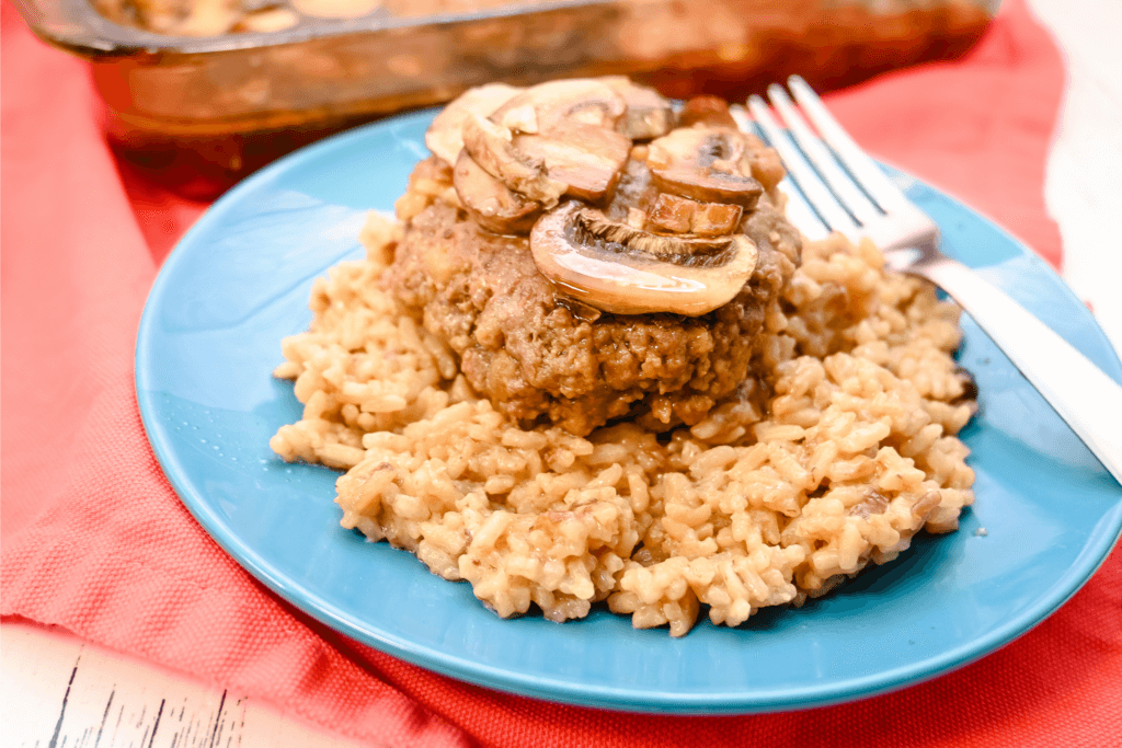 The french onion rice casserole on a blue plate with a salisbury steak on top of it with mushrooms. 