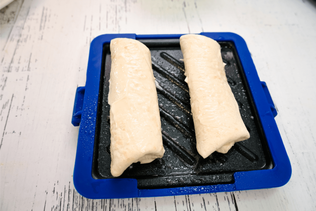 Placing the rolled up chimichangas on the blue microwave toaster which has been sprayed with non-stick oil spray. 