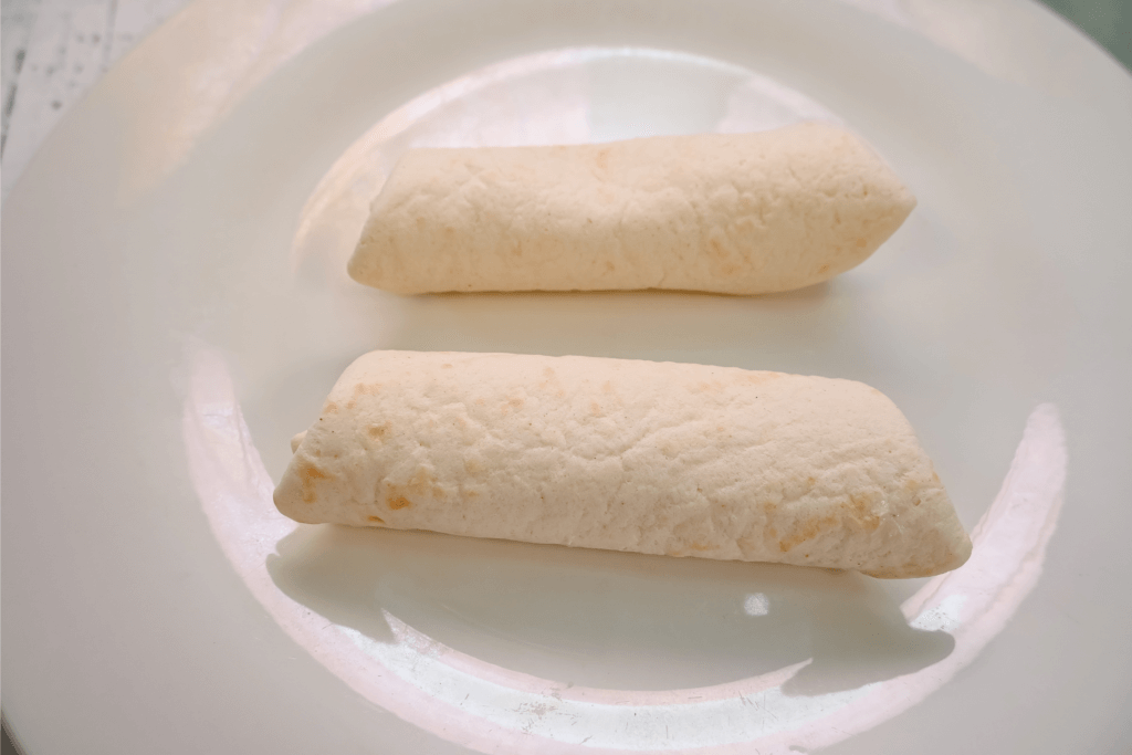 Two flour tortillas with cream cheese filling rolled up on a white plate. 
