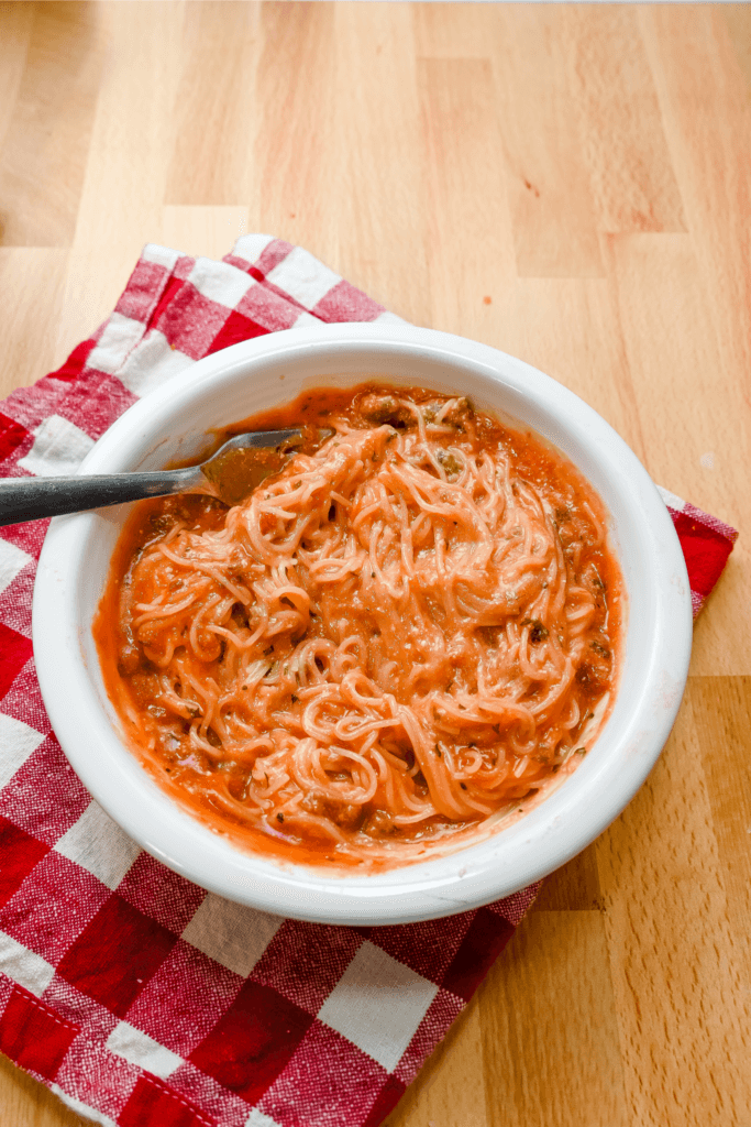 A white bowl on a checked napkin with the cheesy spaghetti all mixed up before adding cheese with a fork in the bowl. 