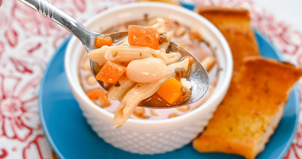 A close up of a spoonful of pasta fagioli soup over a white bowl on a blue plate with garlic toast.  