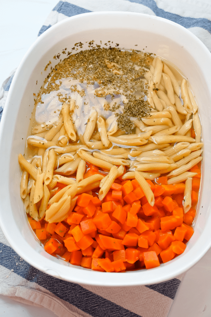 The pasta, carrots, seasoning, and chicken broth in a white microwave-safe container for the pasta fagioli
