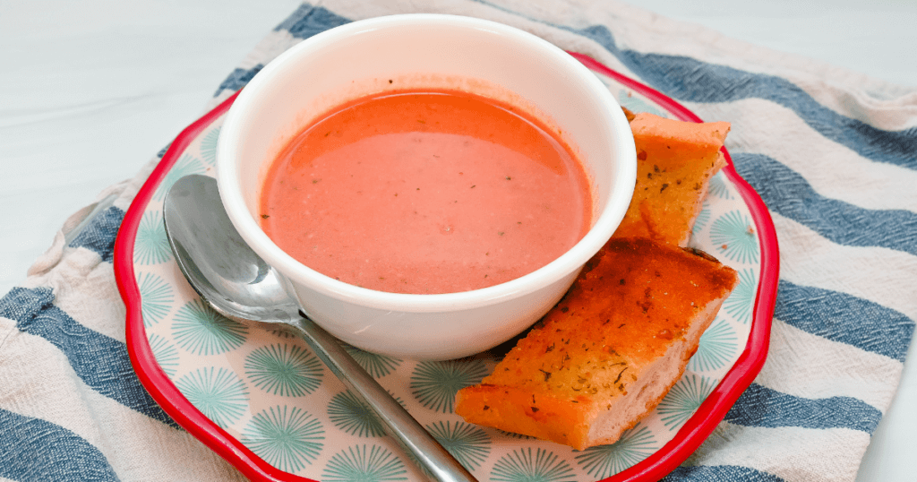 Close up of a bowl of creamy tomato soup on a red rimmed plate with a spoon and garlic toast. 