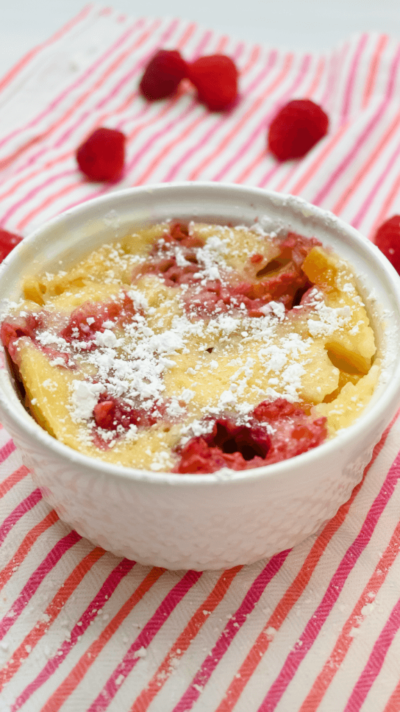 The cooked Berry Peach Clafoutis topped with powdered sugar surrounded by raspberries on a pink striped towel